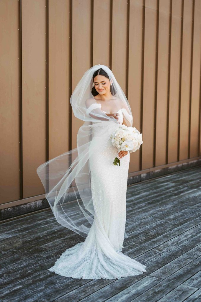 The bride gracefully lifts her veil as it flows in the wind, holding a bouquet of white flowers.