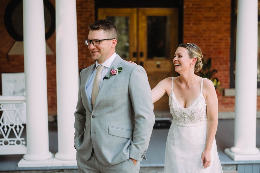 A bride in a lace wedding gown playfully reaching out to tap her groom’s shoulder for their first look.