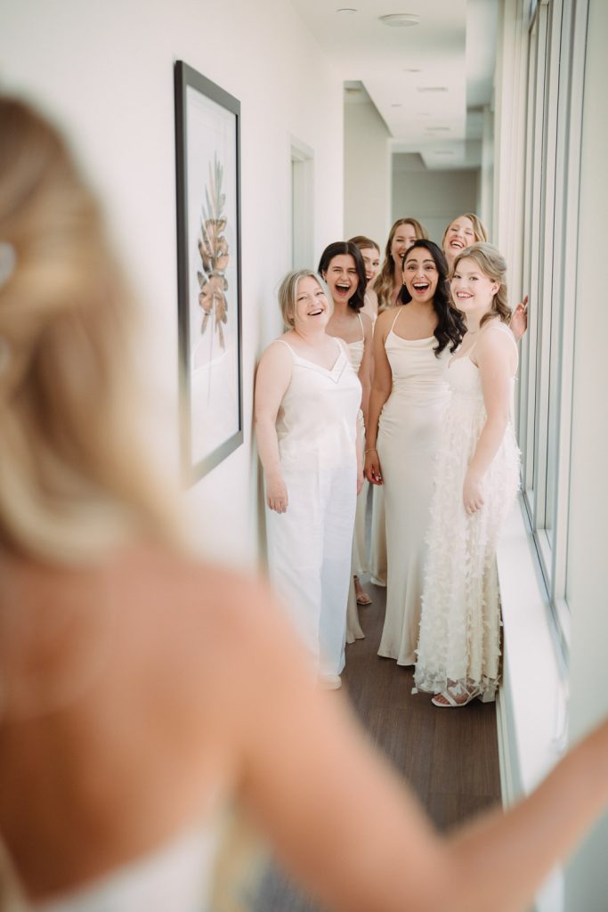 The bridesmaids, dressed in elegant champagne-colored gowns, react with joy and excitement as they see the bride for the first time.